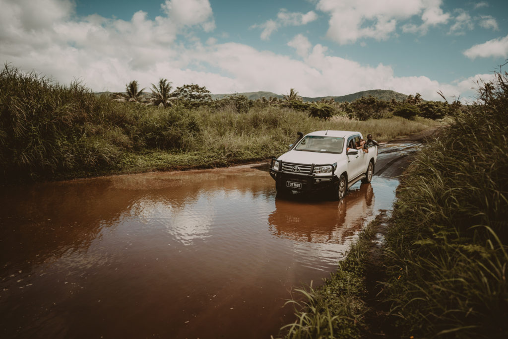 A couple is driving a white truck over a puddle on a dirt road, on their way to get married in Vanuatu.