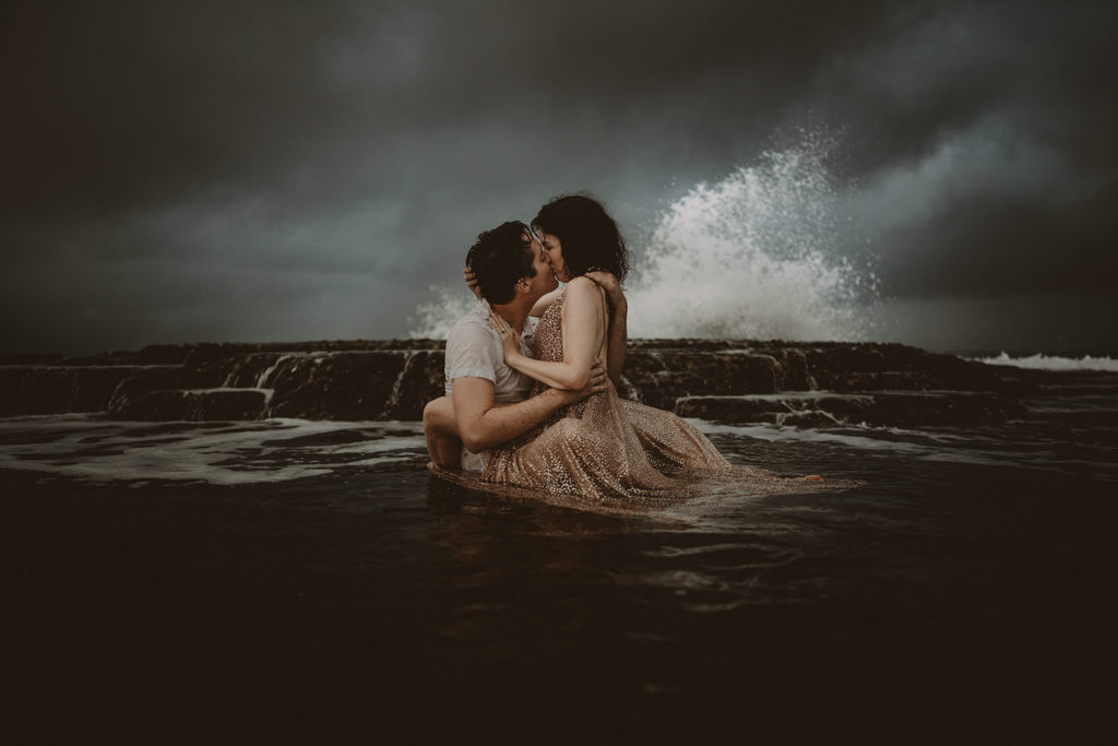 couple kissing after a beach elopement, sitting in the water against a stormy backdrop