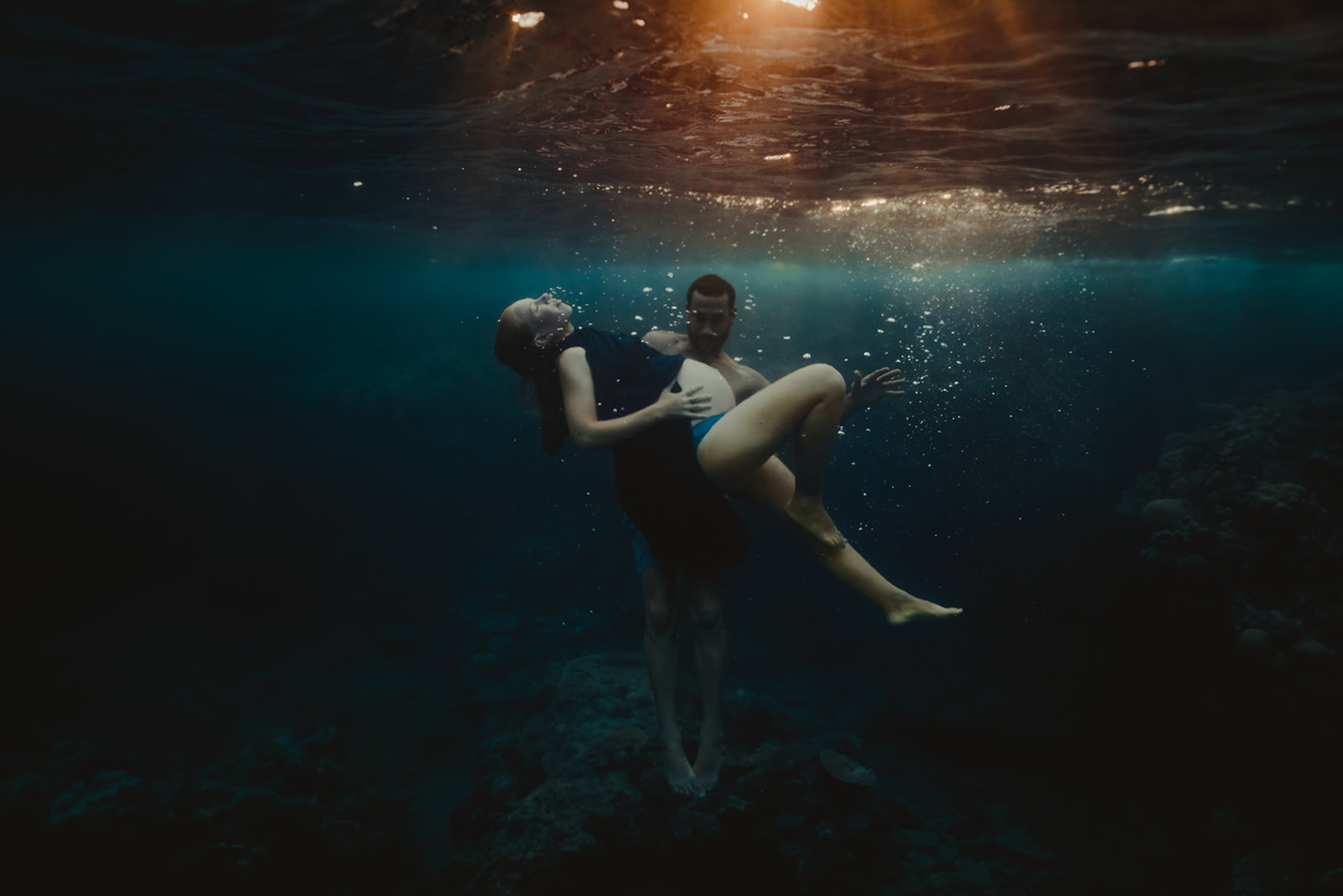 pregnant woman with her partner floating underwater with sunrays breaking through the water.