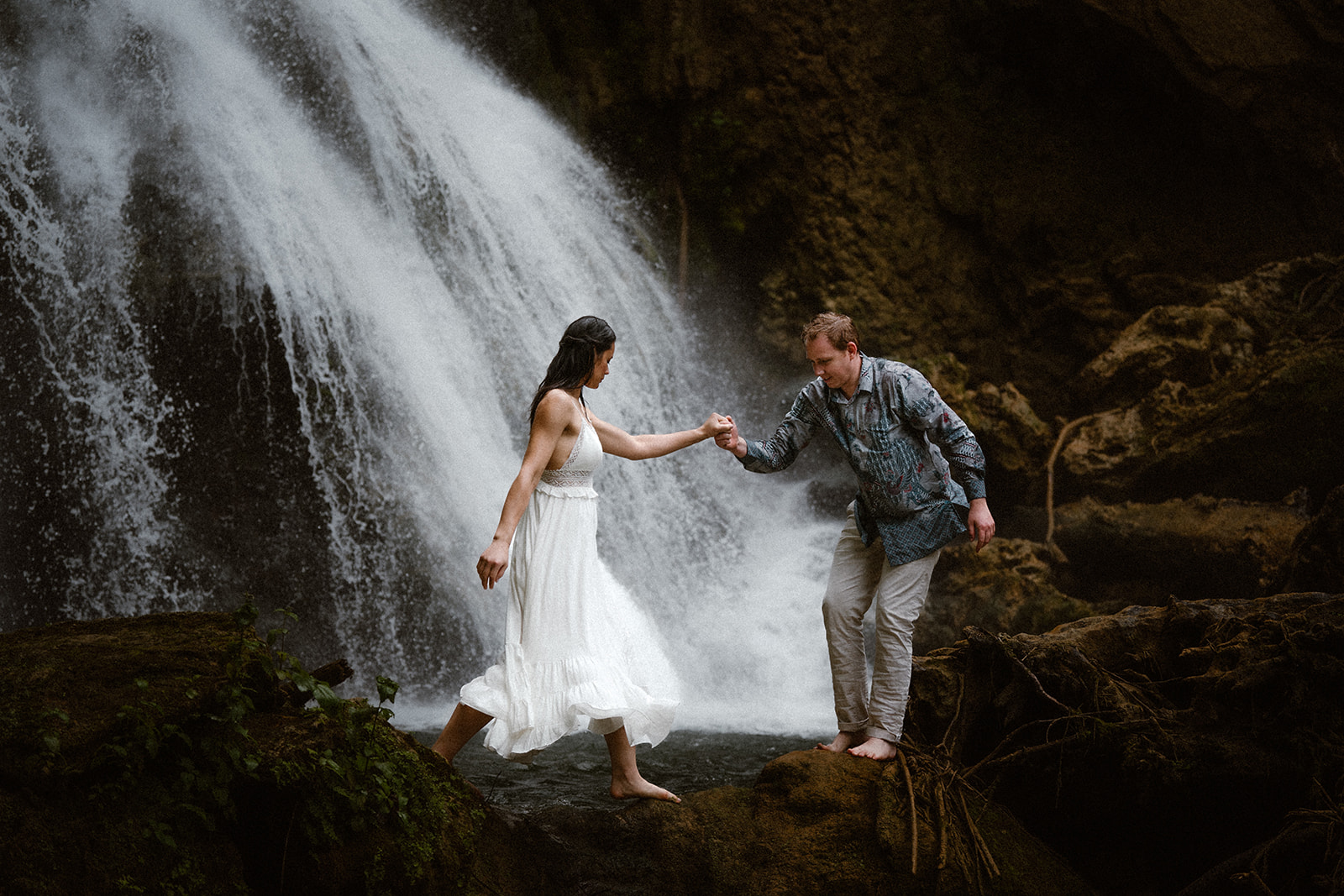 Couple reaching out for each other across a stream, in front of a waterfall, during their pre-wedding photoshoot in Vanuatu